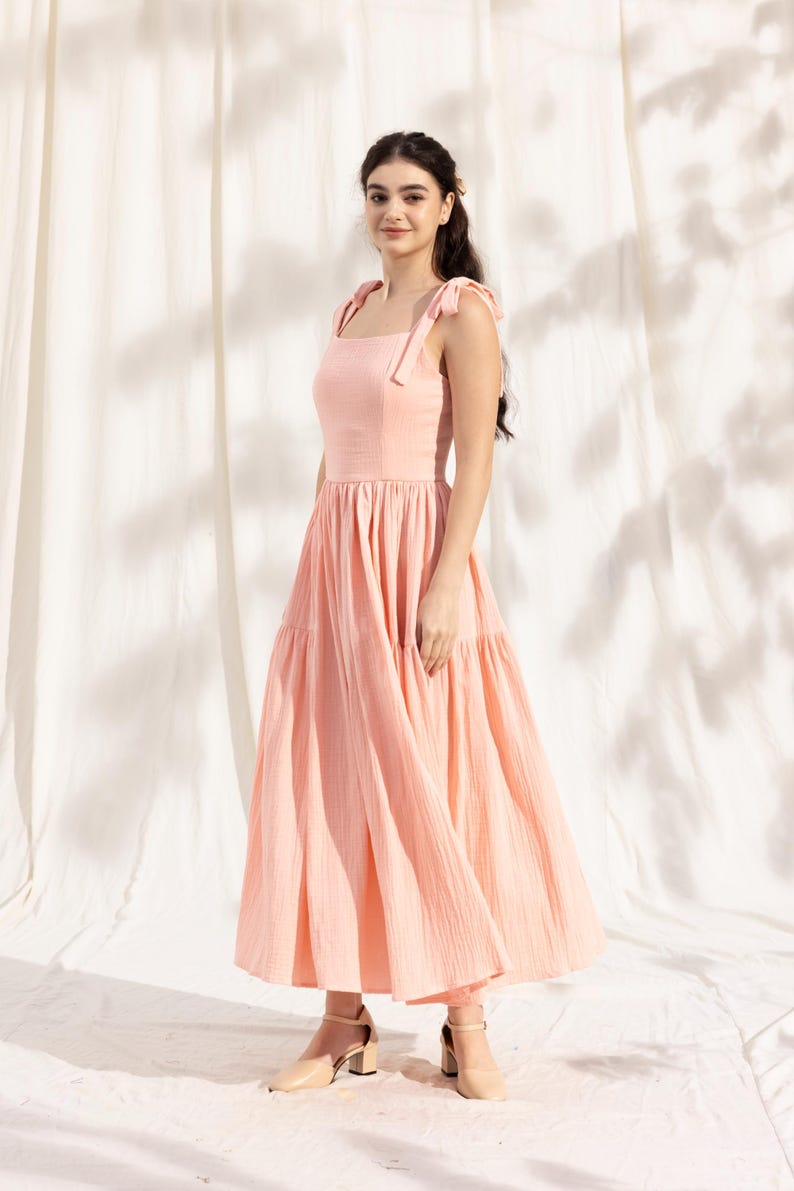 May include: A peach-colored tiered maxi dress with thin straps that tie at the shoulders. The dress is made of a lightweight fabric and has a flowy silhouette.