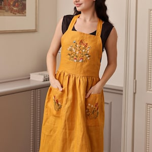 Puede incluir: Un delantal de lino amarillo mostaza con flores bordadas y bolsillos. El delantal tiene una falda fruncida y tirantes ajustables.