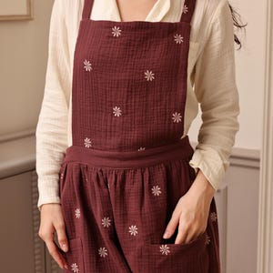 May include: A burgundy apron with white floral embroidery. The apron has a bib, waist tie, and two front pockets. The model is wearing a cream-colored long-sleeved shirt underneath the apron.