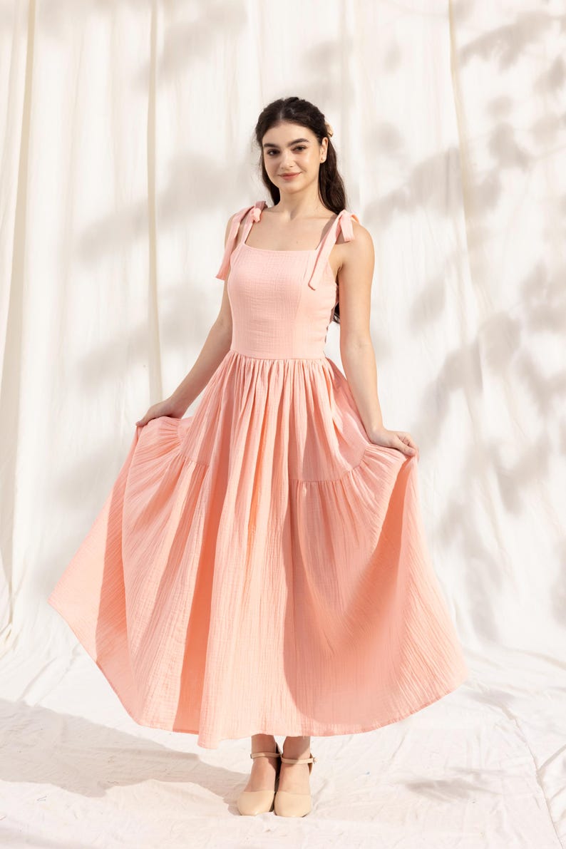 May include: A light pink tiered maxi dress with thin straps that tie at the shoulders. The dress has a square neckline and a flowy skirt.