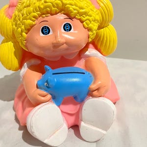 May include: A vintage plastic piggy bank shaped like a girl with blonde hair, wearing a pink dress and holding a blue piggy bank.