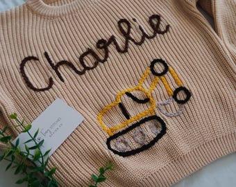 May include: A beige knitted jumper with a brown embroidered name "Charlie" and a yellow embroidered construction vehicle. The jumper has a ribbed texture. A white tag with the text "Tiny Stitches Design Co." is visible in the image.