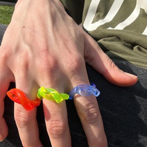 以下が含まれることがあります： 赤、ネオンイエロー、青の3色のカラフルな指輪が指に飾られています。リングはねじれた抽象的なデザインです。手はぼやけた背景の前にあり、屋外の環境を示唆しています。