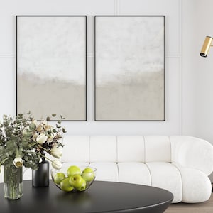 May include: A white sofa with a tufted design sits in a living room with two framed abstract paintings, a vase of white flowers, a bowl of green apples, and a black floor lamp with a gold accent.