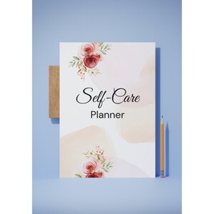 Könnte beinhalten: Ein weißer Self-Care-Planer mit floralen Akzenten und dem Titel in eleganter Schrift. Der Planer hat aquarellartige, pfirsichfarbene Flecken und wird von einem Bleistift und einem Holzblock auf blauem Hintergrund begleitet.