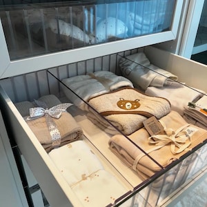 Può includere: Un cassetto bianco con divisori trasparenti è aperto, rivelando una varietà di vestiti e asciugamani per bambini piegati. Il cassetto è organizzato con diversi articoli in ogni sezione. Gli articoli includono un asciugamano bianco con un disegno di orso marrone, un asciugamano beige, una coperta bianca con un motivo marrone e una coperta beige con un nastro marrone.