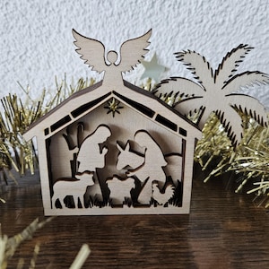 May include: A wooden nativity scene with an angel on top, a star, and a palm tree. The scene depicts Mary, Joseph, and baby Jesus in a stable with animals. Gold tinsel and star decorations surround the scene.