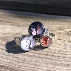Puede incluir: Tres anillos de plata con diferentes imágenes en la parte superior. El primer anillo tiene una imagen en blanco y negro de una cara. El segundo anillo tiene una imagen en blanco y negro de dos manos. El tercer anillo tiene una imagen roja y blanca de una cara.