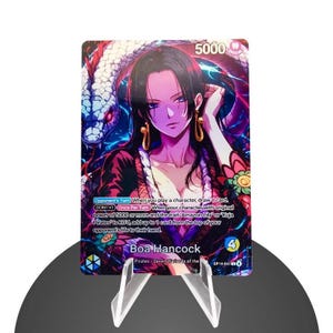 Può includere: Una carta collezionabile con Boa Hancock della serie "One Piece". La carta mostra una vivace illustrazione del personaggio con un serpente e il testo "5000". La carta è esposta su un supporto di plastica trasparente.