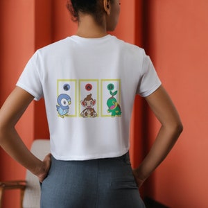 May include: White cropped t-shirt with a graphic of three cartoon characters in yellow frames. The characters are a blue bird, a brown monkey, and a green turtle.