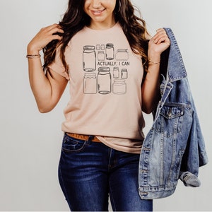 May include: A light pink t-shirt with a graphic of multiple mason jars and the text "Actually, I can".