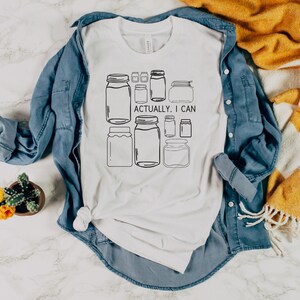 May include: White t-shirt with a graphic of jars and the text "Actually, I can". The t-shirt is folded and laid on a blue denim shirt and a white and yellow striped blanket.