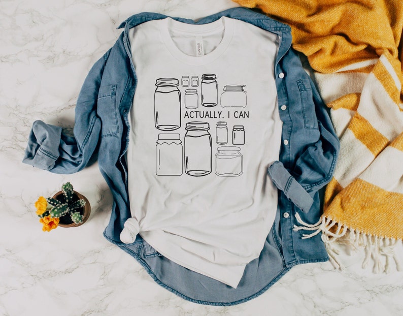 May include: White t-shirt with a graphic of jars and the text "Actually, I can". The t-shirt is folded and laid on a blue denim shirt and a white and yellow striped blanket.