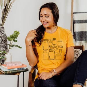 May include: A woman wearing a yellow t-shirt with a graphic of mason jars and the text "Actually, I can".