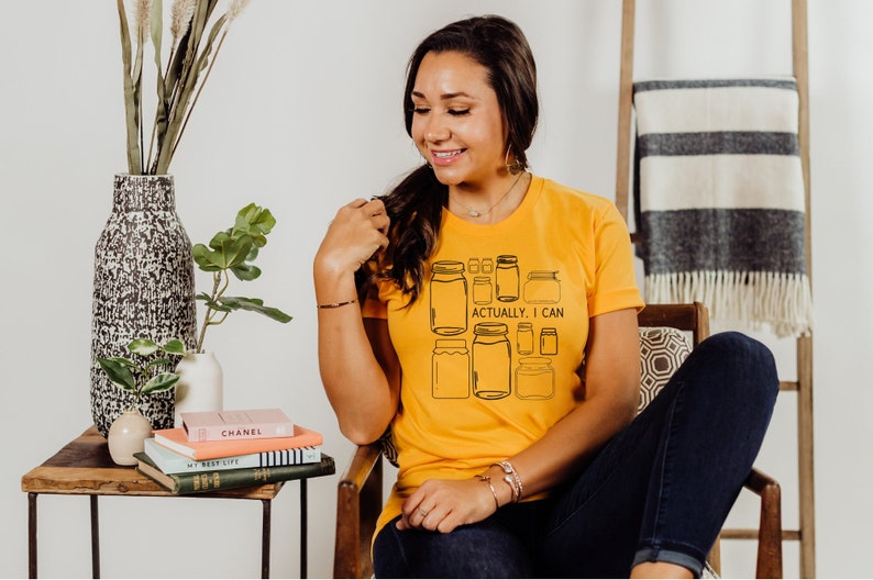 May include: A woman wearing a yellow t-shirt with a graphic of mason jars and the text "Actually, I can".