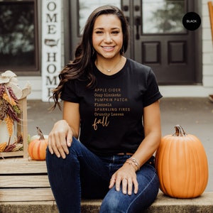 Puede incluir: Camiseta negra con las palabras "APPLE CIDER, Cozy blankets, PUMPKIN PATCH, Flannels, SPARKLING FIRES, Crunchy Leaves, fall" en color cobre. La persona lleva vaqueros azules y está sentada junto a una calabaza naranja.