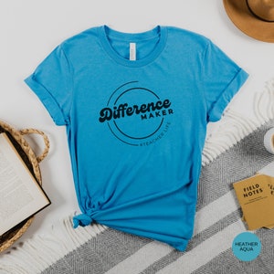Op de afbeelding: Een turquoise blauw t-shirt met een zwarte cirkelvormige afbeelding met de tekst "Difference Maker #Teacher Life".
