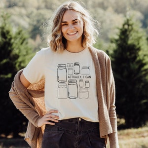 May include: A white t-shirt with a graphic of multiple mason jars and the text "Actually, I can". The person wearing the shirt is standing in front of a green background.
