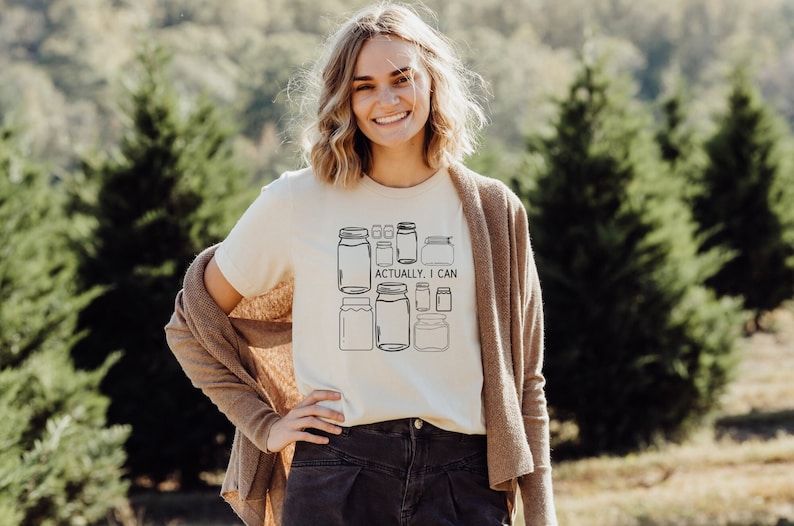 May include: A white t-shirt with a graphic of multiple mason jars and the text "Actually, I can". The person wearing the shirt is standing in front of a green background.