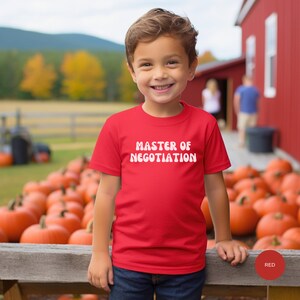 May include: A red t-shirt with white text that says "Master of Negotiation". The shirt is being worn by a young boy standing in front of a pile of pumpkins.