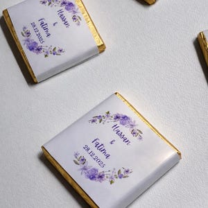 May include: Square chocolate bars with white wrappers and gold foil edges. Each wrapper is decorated with a purple floral design and the names "Hassan & Fatima" with the date "28.12.2025".