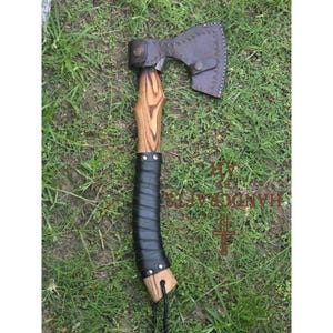 Könnte beinhalten: Axt mit Holzgriff und dunkler Metallklinge. Der Griff ist mit schwarzem Leder umwickelt. Eine braune Lederscheide bedeckt die Klinge. Die Axt liegt auf grünem Gras.