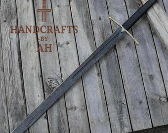 May include: A long steel sword with a black handle and gold crossguard, lying on a wooden surface. The blade has a detailed pattern. The text "HANDCRAFTS BY AH" is visible.