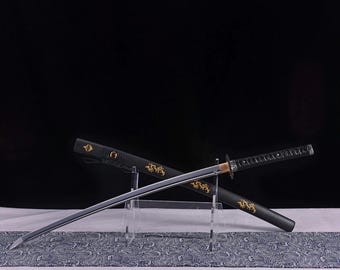 May include: A Japanese katana sword with a silver blade and black handle, resting on a clear stand. The black scabbard features gold dragon designs. The sword is displayed against a dark background.