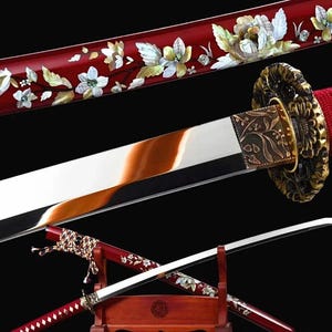 Op de afbeelding: Een Japanse katana-zwaard met een rood gelakte schede en handvat, versierd met bloemige parelmoer inlegwerk. Het lemmet is van gepolijst staal met een zichtbare temperlijn. De tsuba heeft een gedetailleerd bronzen ontwerp.