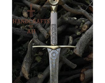 May include: A detailed handcrafted sword. The hilt is white with an ornate pattern, and the crossguard and pommel are gold-coloured. The blade is silver with etched details. The text "HANDCRAFTS BY AH" is visible.