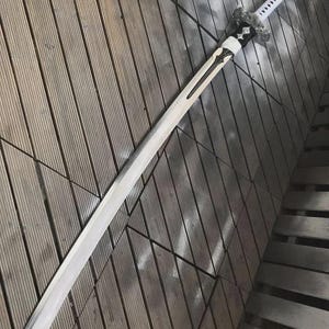 May include: A long, white sword with a silver blade and a white handle. The sword has a decorative guard and a tassel at the end of the handle. The sword is lying on a wooden surface.