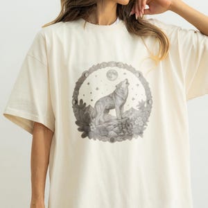 May include: An off-white short-sleeve t-shirt featuring a circular graphic on the chest. The design depicts a wolf howling at a moon surrounded by stars, mountains, and foliage, rendered in a monochromatic gray sketch style on the fabric.