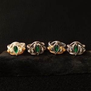 May include: Four silver rings with a snake design, each featuring a green gemstone center. The rings are displayed on a dark surface. Some rings have gold accents on the snake's base.