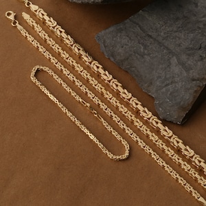 May include: Four gold chain necklaces of varying lengths and widths, displayed on a brown surface. The chains are made of a woven design.