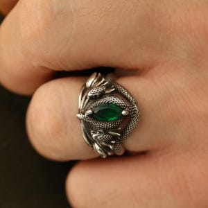 Op de afbeelding: Een zilveren ring met een slangenmotief, met een ovale, smaragdgroene edelsteen. Het ontwerp van de ring omvat getextureerde schubben en de kop en staart van de slang die de groene steen omlijsten.