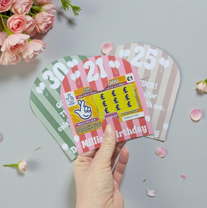 May include: A hand holding a pink and white striped birthday card with a scratch-off lottery ticket. The card is labeled "21" and has text that reads "Millie Birthday". Other cards are visible with the numbers "30" and "25".