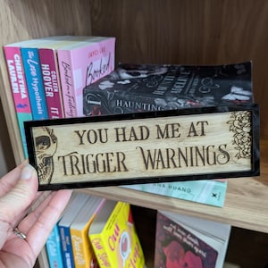 May include: A wooden sign with black trim and a skull design. The sign reads "You had me at trigger warnings".