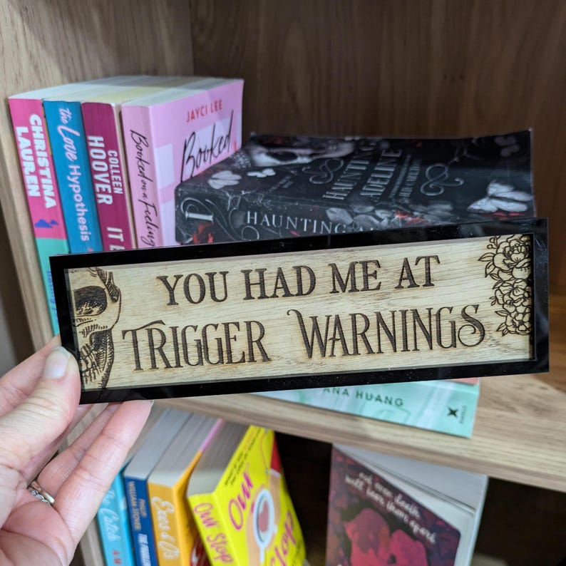 May include: A wooden sign with black trim and a skull design. The sign reads "You had me at trigger warnings".