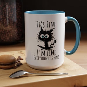 Puede incluir: Taza de cerámica blanca con borde y asa azules. La taza presenta una ilustración en blanco y negro de un gato con el texto "It's Fine I'm Fine Everything is Fine."