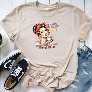 Op de afbeelding: Beige T-shirt met een retro illustratie van een vrouw met een rode polkadot hoofddoek die een kopje koffie vasthoudt. De tekst op het shirt luidt: "OF COURSE SIZE MATTERS, NO ONE WANTS A SMALL CUP OF COFFEE". Zwarte en witte sneakers en een blauwe jeans zijn ook zichtbaar.