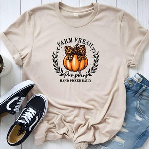 Könnte beinhalten: Beiges T-Shirt mit einem Kürbis-Motiv, verziert mit einer Leopardenmuster-Schleife, umgeben von einem Kranz. Der Text auf dem Shirt lautet "FARM FRESH PUMPKINS HAND PICKED DAILY". Schwarz-weiße Turnschuhe und blaue Jeans sind ebenfalls im Bild.