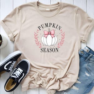 Könnte beinhalten: Beiges T-Shirt mit einem weißen Kürbis-Design mit rosa Schleife und Kranz, sowie dem Schriftzug "PUMPKIN SEASON". Auf dem Bild sind auch schwarz-weiße Turnschuhe und blaue Jeans zu sehen.
