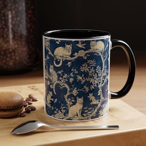 May include: A ceramic mug with a black handle and interior. The mug features a dark blue background with a repeating pattern of cats perched on branches with leaves and flowers in a cream color. A spoon and a pastry are in the foreground.