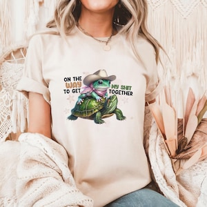 May include: Beige t-shirt with a humorous graphic of a frog in a cowboy hat riding a turtle. The text reads "ON THE WAY TO GET MY SHIT TOGETHER". The design is playful and colourful.