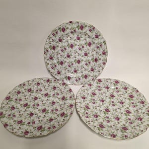 May include: Three white ceramic plates with a floral pattern of small purple roses and green leaves. The plates have a scalloped edge with a gold trim. The plates are arranged in a triangular shape.