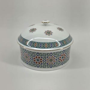 May include: A white ceramic lidded serving dish with a gold rim and intricate geometric patterns in green, blue, and rust. The lid features small floral designs. A small gold knob adorns the top of the lid.