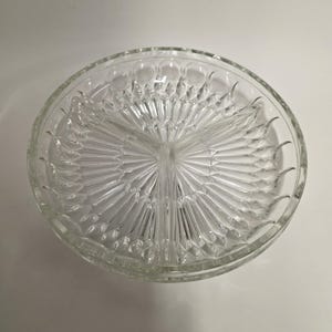 May include: A clear, round, divided glass serving dish. The dish is segmented into three sections, with a central hub and radiating lines. The rim features a pattern of small, raised circles. Ideal for serving snacks or appetizers.