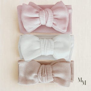 Könnte beinhalten: Drei Haarband-Sets in Pastellfarben mit Waffelstruktur: Rosa, Weiß und Beige. Jedes Haarband hat eine große dekorative Schleife. Die Haarbänder sind auf einem hellbeigen Hintergrund angeordnet.