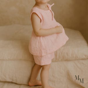 May include: A light pink toddler outfit, consisting of a top and shorts. The top has a round neckline and a loose, flowing fit. The shorts are a matching light pink color. The outfit is made of a soft, textured fabric.