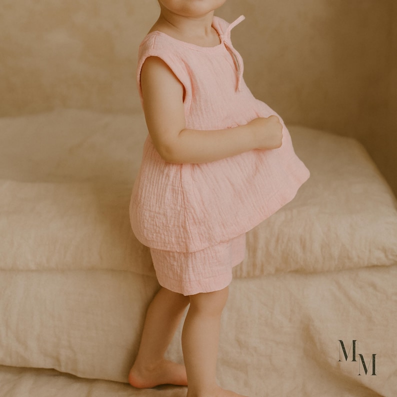 May include: A light pink toddler outfit, consisting of a top and shorts. The top has a round neckline and a loose, flowing fit. The shorts are a matching light pink color. The outfit is made of a soft, textured fabric.